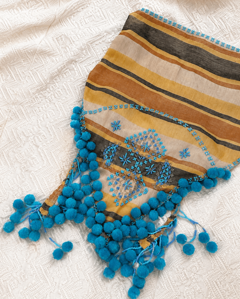 Zarah Juan bupanda scarf with turquoise beadwork and pom-pom trim laid on a textured ivory fabric.